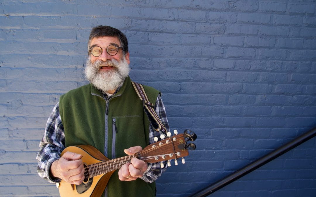 Mand with a beard and glasses smiling and strumming the mandolin
