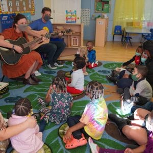 Two music teachers playing guitar in front of a group of young children sitting on a carpet.