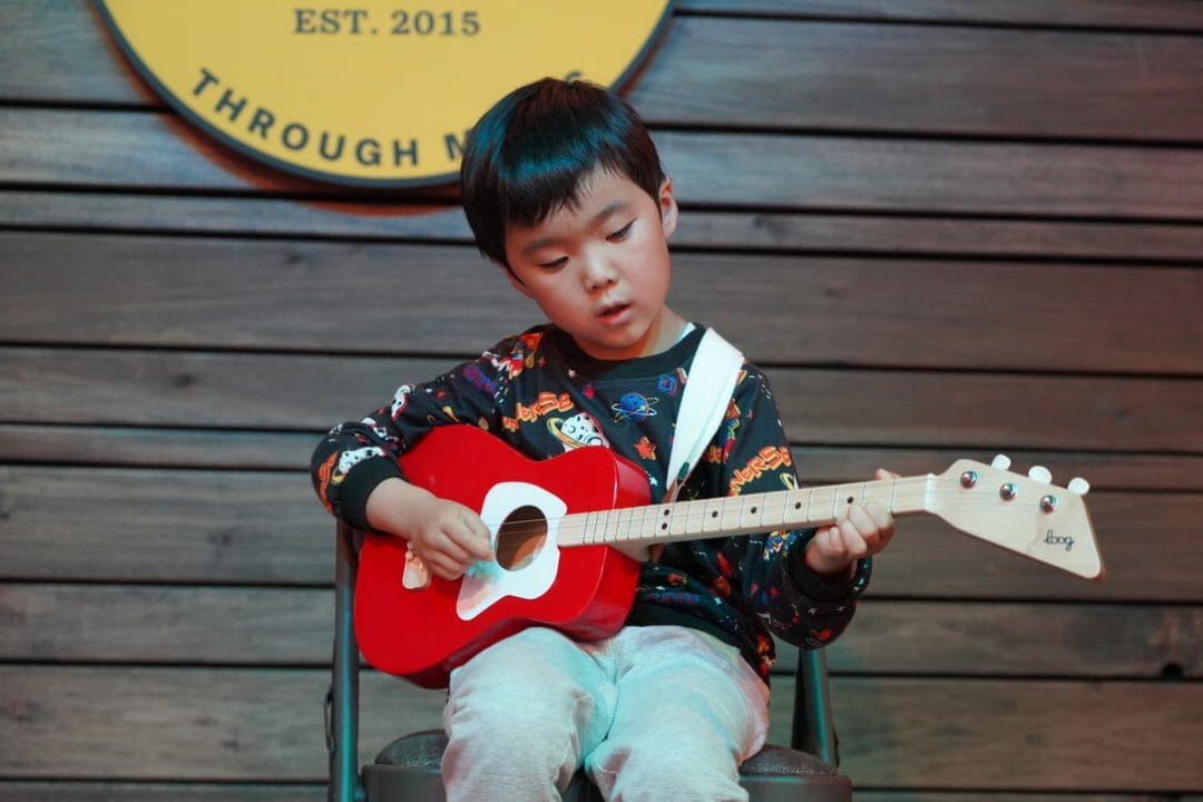 6 year old boy sitting in a folding chair with grey sweatpants and a black sweatshirt playing a 3 string bright red guitar. 
